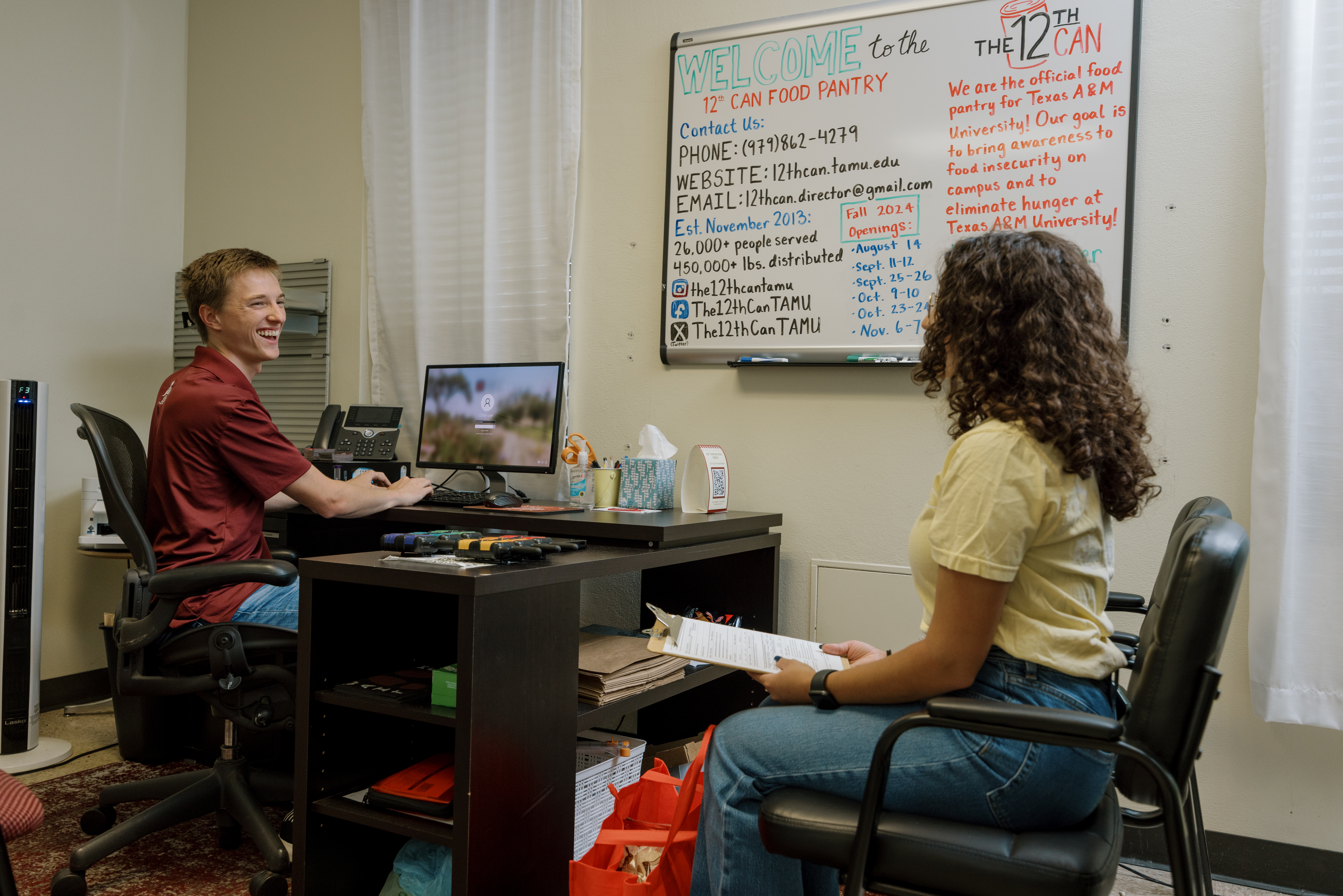 Two 12th Can members talking across a desk, one at a computer and the other holding a clipboard.